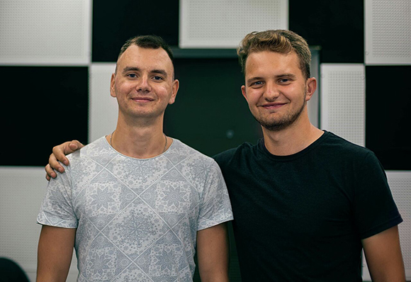 Two smiling men standing side by side with arms around each other against a black and white checkered wall.
