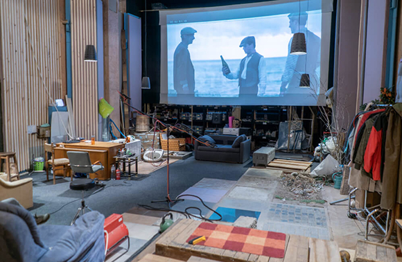A film set featuring a large projection screen, studio lighting, cameras, and various production equipment.