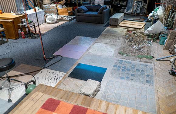 A room featuring mismatched floor tiles, a dark sofa, a wooden desk, and various construction materials.