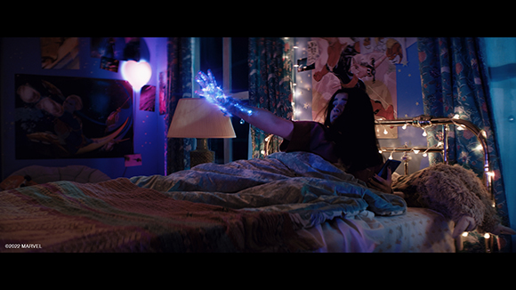 A person lying in bed reaches out a hand glowing with bright blue magical energy in a dimly lit room.