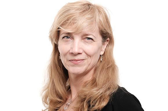 A woman with wavy blonde hair and silver earrings smiles slightly against a white background.