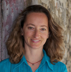 A woman with shoulder-length wavy brown hair and a blue collared shirt smiles forward. She is standing in front of a stone wa