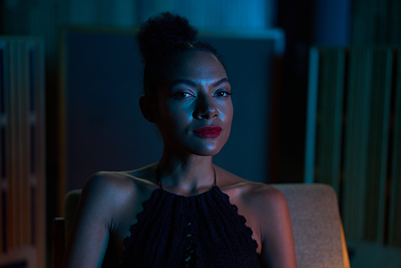 A woman with dark hair in a bun looks forward under dramatic blue and red lighting.