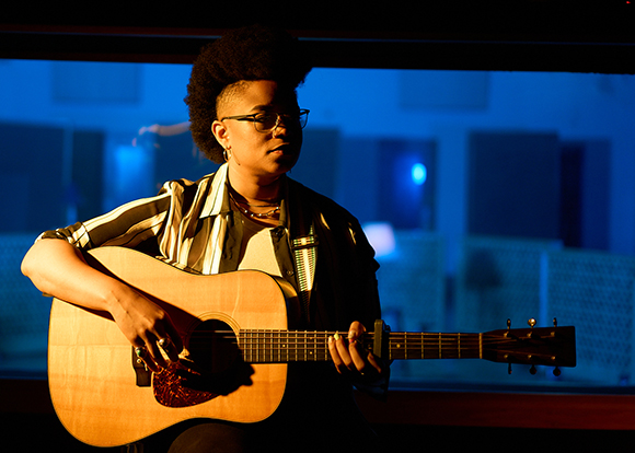 A person with an afro and glasses playing an acoustic guitar under blue light.