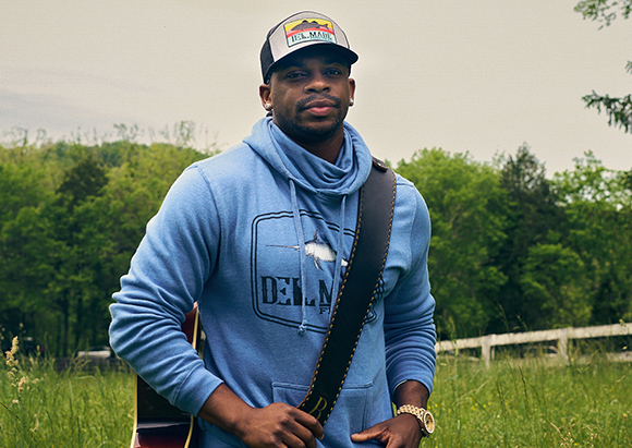 A man wearing a blue hoodie and a baseball cap stands in a grassy field with a guitar strap over his shoulder.