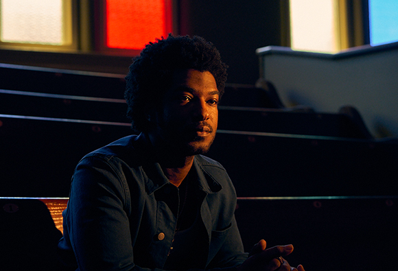 A man with dark curly hair sits in a dark theater seat, looking off to the side.