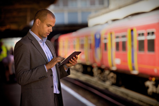 A man in a grey suit stands on a platform using a digital tablet with a red train blurred in the background.