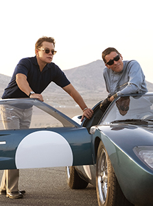 Two men in sunglasses lean over a dark green vintage sports car in a desert landscape.