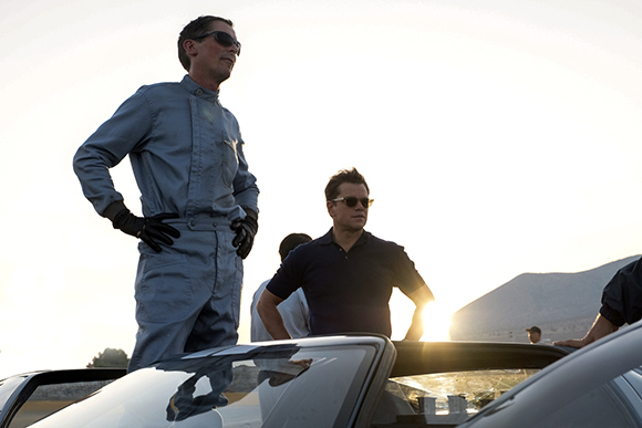 A man in blue coveralls and sunglasses stands by a car next to a man in a dark polo shirt.
