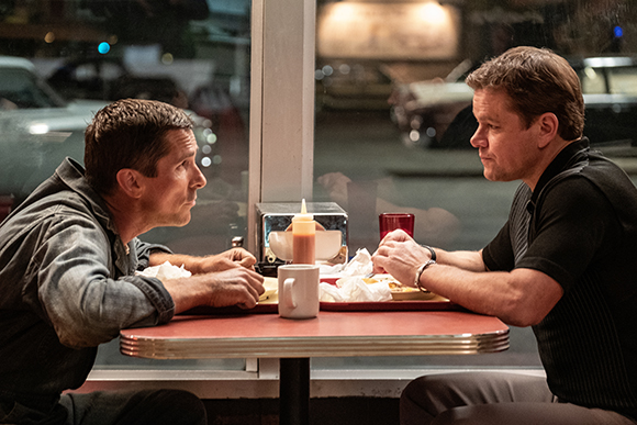 Two men sit facing each other at a small table inside a diner at night.