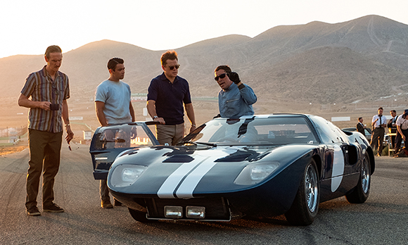 Four men stand near a blue vintage racing car with white stripes on a track against a mountain backdrop.
