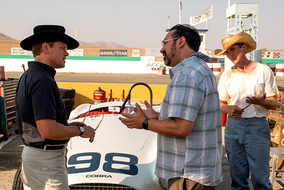 Three men in casual attire stand near a white Cobra race car at a racetrack.