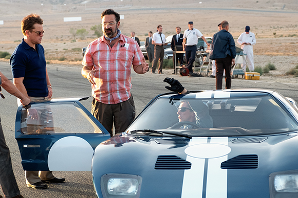 A man in a plaid shirt gestures toward a man in a blue polo standing by a blue racing car on a track.