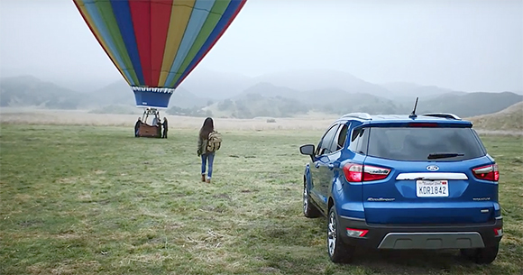 A blue Ford SUV parked in a grassy field near a colorful hot air balloon and a person walking away.
