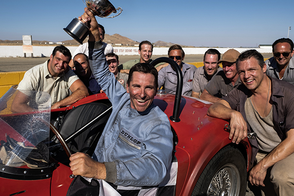 A man in a blue racing suit holds a trophy while sitting in a red vintage race car, surrounded by cheering men.