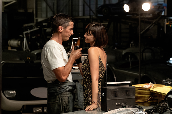 A man in a white t-shirt and a woman in a patterned dress stand close together in a dark garage.
