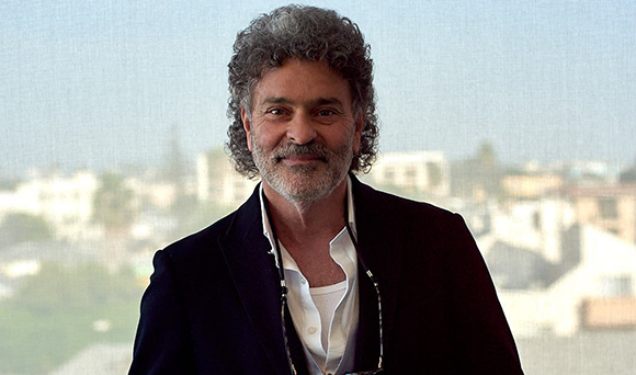 A man with curly grey hair and a beard wearing a dark blazer and white shirt.