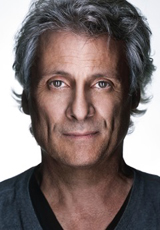 A middle-aged man with wavy gray hair and light eyes looking directly at the camera against a white background.