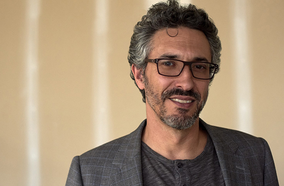 A man with curly graying hair and glasses wearing a gray blazer and dark shirt.