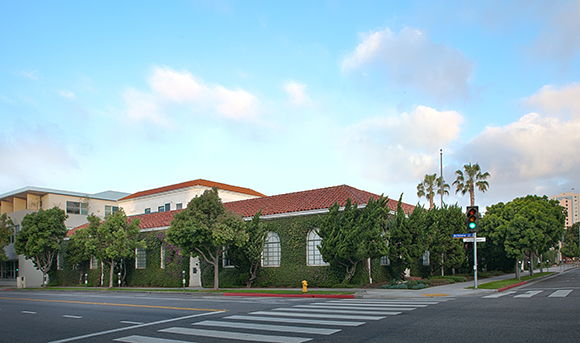 A building with a red tile roof covered in green ivy sits behind trees at a street intersection.