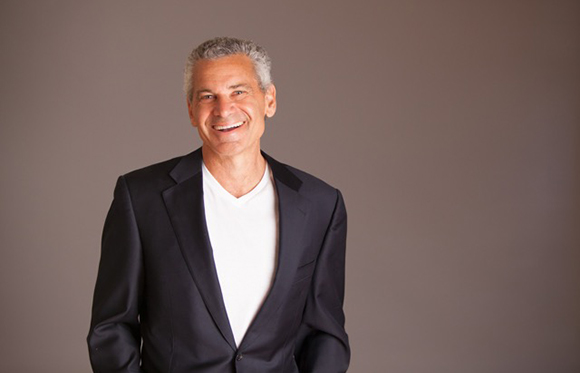 A smiling man with graying hair wearing a dark blazer over a white v-neck t-shirt against a solid background.