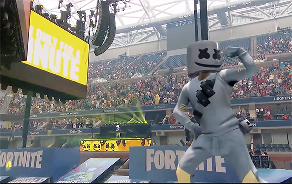 A person in a grey Marshmello costume dancing in a crowded sports stadium.