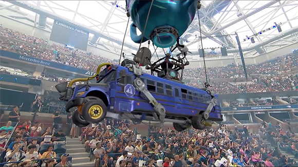 A blue bus hangs from a large teal balloon over a packed sports stadium crowd.