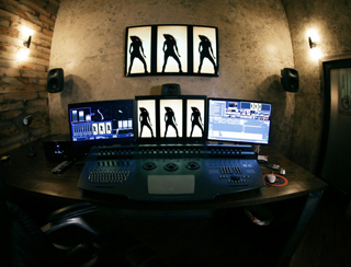 A desk with three computer monitors, a large mixing console, and studio speakers in a dimly lit room.