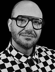 Black and white portrait of a man with glasses and a checkered shirt against a black background.