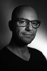 Black and white close-up portrait of a bald man wearing dark-rimmed glasses.