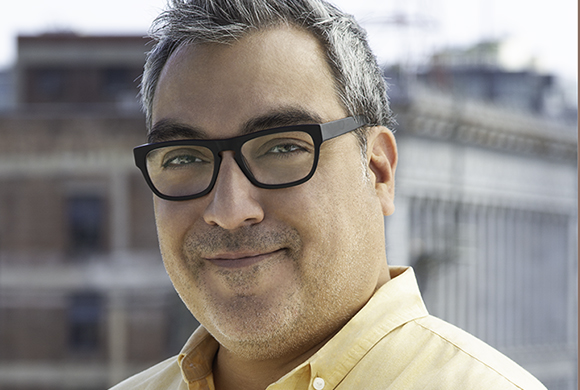 A man with salt-and-pepper hair and black-rimmed glasses smiling in front of a blurred city background.