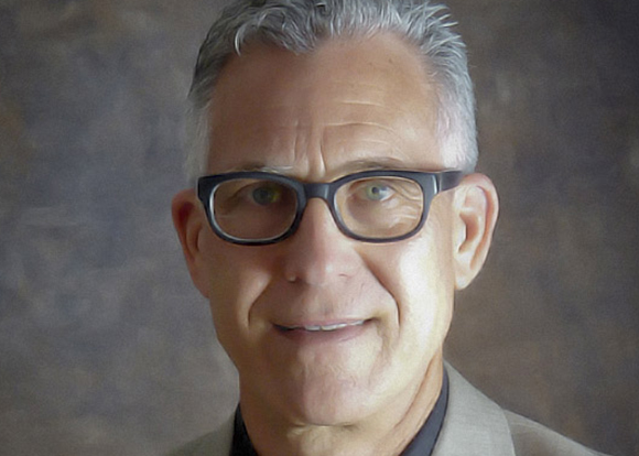 A man with short grey hair and black-rimmed glasses smiling against a dark, textured background.