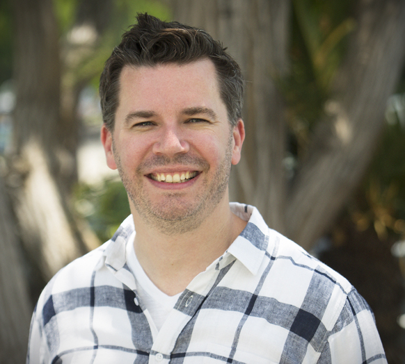 A man with short dark hair and light stubble smiling while wearing a white and navy plaid shirt.