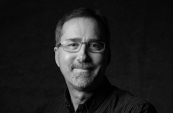 Black and white headshot of a middle-aged man with glasses and a goatee smiling against a dark background.