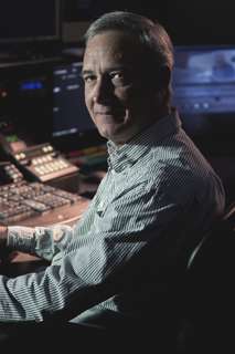 A man in a striped shirt sits before a professional audio or video mixing console in a dimly lit studio.