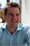 A smiling man with short brown hair wearing a light blue collared shirt.