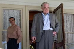 A man in a grey suit stands in a doorway while another man in a tan shirt stands behind him.
