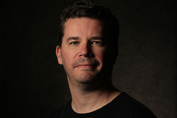 A man with dark wavy hair and light stubble looks forward against a dark, textured background.