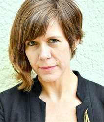 A woman with short brown hair and green eyes looking directly at the camera against a light green wall.
