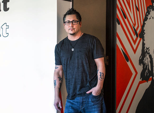 A man with glasses and tattoos wearing a dark grey t-shirt and blue jeans stands in a doorway.