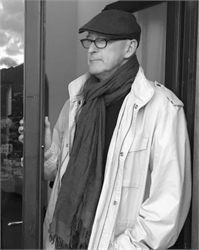 A man wearing a flat cap, glasses, a scarf, and a light jacket stands in a doorway.