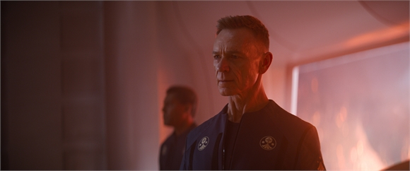 A man in a dark blue uniform with circular patches looks forward in a room with warm, reddish lighting.