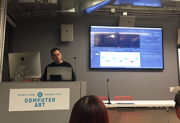A man stands at a podium with a laptop and a computer monitor, presenting in front of a large screen displaying animation sof