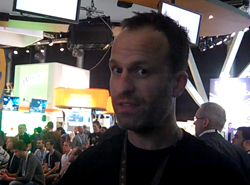 A man with short dark hair looks toward the camera in a brightly lit, crowded convention hall.