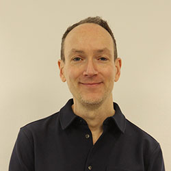 A man with short hair and a slight smile wearing a dark navy button-down shirt.