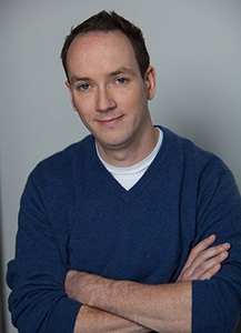 A man with short brown hair and a light complexion wears a blue v-neck sweater over a white t-shirt. He has his arms crossed