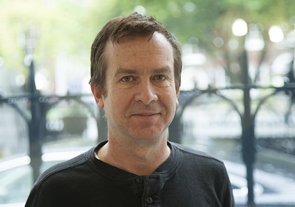 A man with short brown hair and a black shirt smiling at the camera in an outdoor setting.