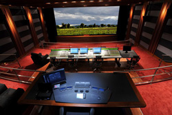 A wide view of a dark screening room featuring a large landscape screen, mixing desks, and red carpeting.