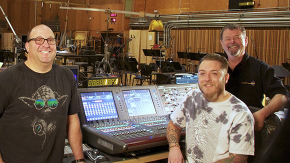 Three men smiling behind a large Yamaha audio mixing console in a professional recording studio.