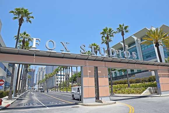 Large silver letters spelling FOX STUDIOS atop a wide entrance gate under a clear blue sky.